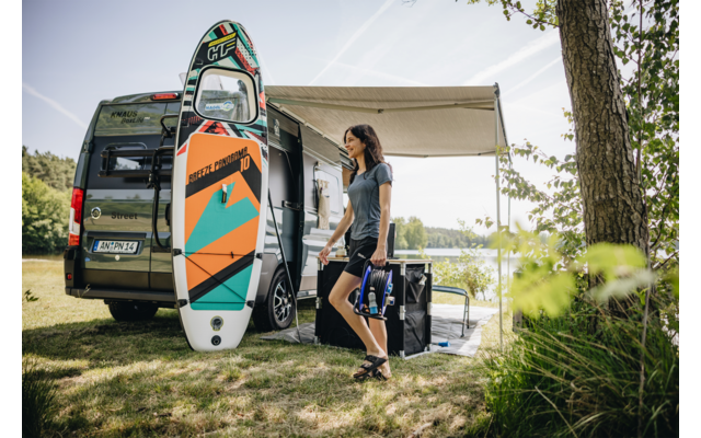 Frau steht neben offenem Campingbus mit ausgefahrener Markise, buntes Stand-up-Paddle-Board lehnt am Bus, See und Bäume im Hintergrund