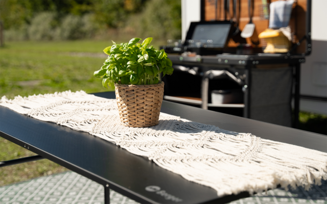 Geflochtener Korb mit grüner Topfpflanze auf weißem Makramee-Läufer auf schwarzem Campingtisch; im Hintergrund ein Grill und eine Wiese