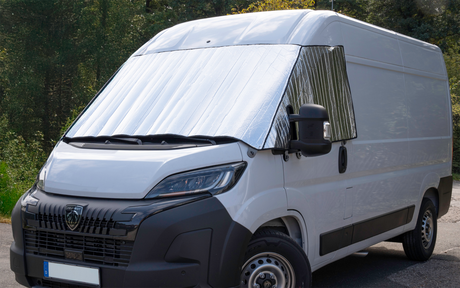 Weißer Kastenwagen in drei-von-vorne-Ansicht mit silbernem Sonnenschutz in Windschutzscheibe und seitlichem Fenster, schwarze Frontpartie und Spiegel, Rad sichtbar, Bäume im Hintergrund