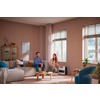 Zwei Personen in einem Wohnzimmer: eine sitzt auf einem grauen Sofa, die andere kniet neben einem kleinen weißen Gerät vor zwei Fenstern mit Jalousien; Couchtisch, Kissen, Teppich und ein Schrank mit Deko sichtbar