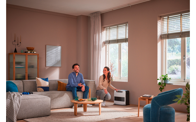 Zwei Personen in einem Wohnzimmer: eine sitzt auf einem grauen Sofa, die andere kniet neben einem kleinen weißen Gerät vor zwei Fenstern mit Jalousien; Couchtisch, Kissen, Teppich und ein Schrank mit Deko sichtbar