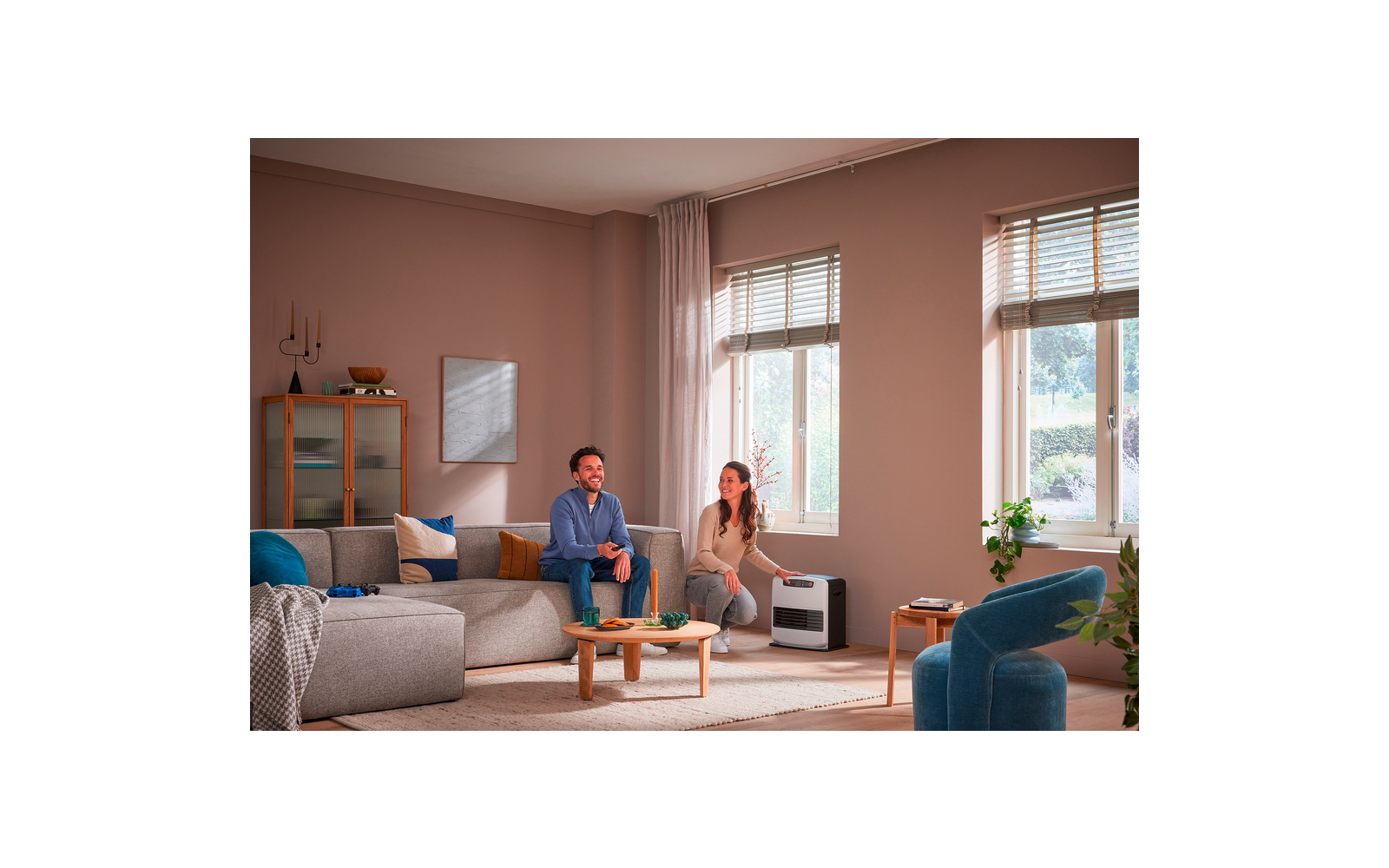 Zwei Personen in einem Wohnzimmer: eine sitzt auf einem grauen Sofa, die andere kniet neben einem kleinen weißen Gerät vor zwei Fenstern mit Jalousien; Couchtisch, Kissen, Teppich und ein Schrank mit Deko sichtbar