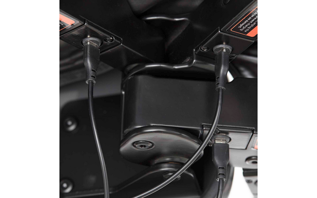 Unterseite eines schwarzen Sitzgestells mit zwei runden Steckern und Kabeln, einem USB-Kabel, Schrauben und orangefarbenen Warnaufklebern