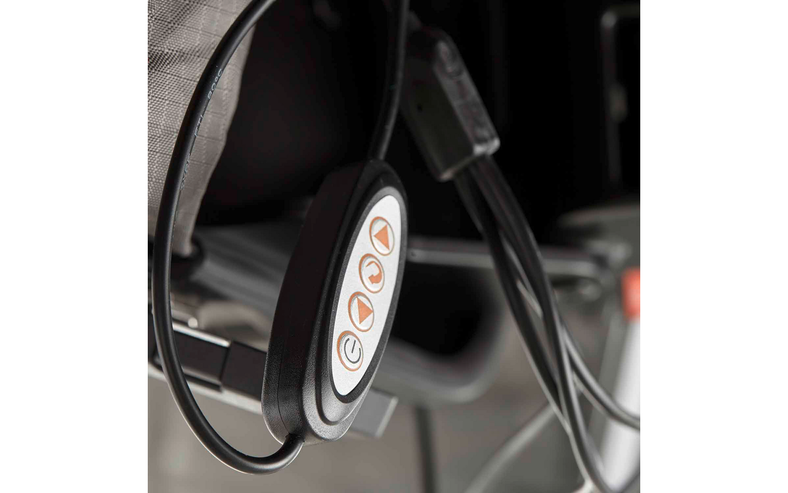 Schwarze ovale Fernbedienung mit drei orangefarbigen Symboltasten auf weißer Einfassung, an Kabeln hängend; unscharfer Stoffbezug und Metallgestell im Hintergrund