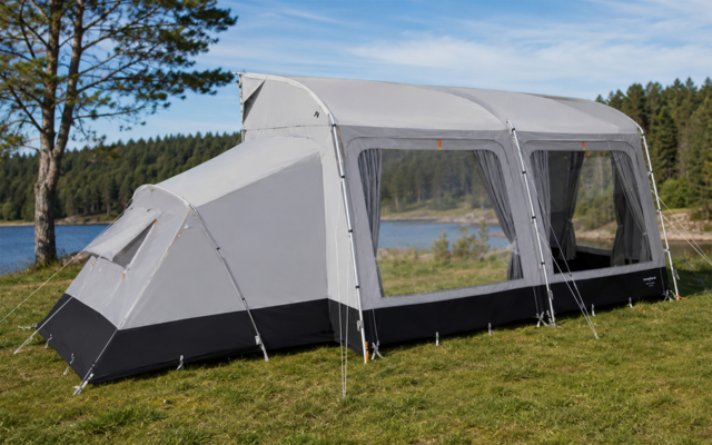 Grote grijs-zwarte tent met een voorbouw en drie grote ramen, opgezet op gras bij een meer; bomen en blauwe lucht op de achtergrond