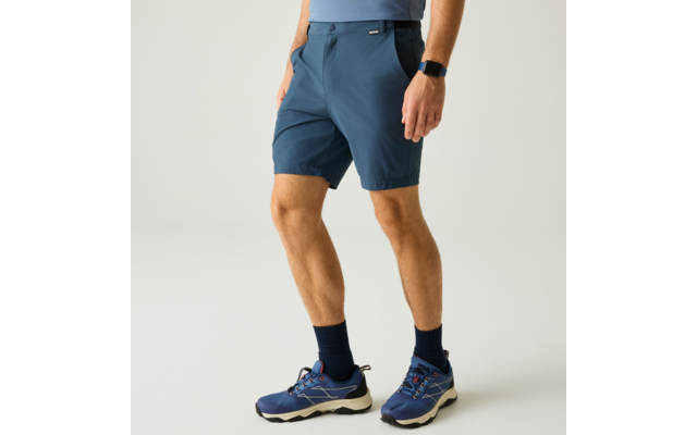 Unterkörper einer Person mit blauen Shorts, dunklen Socken, blauen Sportschuhen mit weißer Sohle und Smartwatch am linken Handgelenk auf hellem Hintergrund