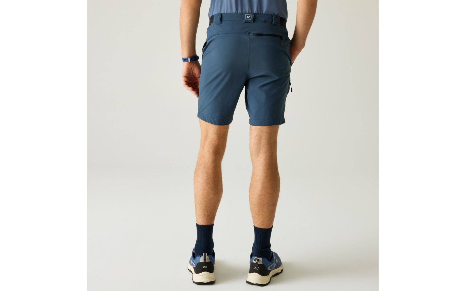Rückansicht einer stehenden Person von der Taille abwärts, dunkelblaue Shorts, hellblaues eingestecktes Shirt, Hände in Gesäßtaschen, dunkle Socken, blaue Sportschuhe mit weißen Sohlen, einfarbiger heller Hintergrund
