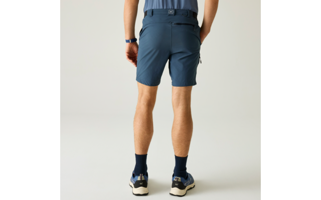 Rückansicht einer stehenden Person von der Taille abwärts, dunkelblaue Shorts, hellblaues eingestecktes Shirt, Hände in Gesäßtaschen, dunkle Socken, blaue Sportschuhe mit weißen Sohlen, einfarbiger heller Hintergrund