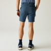 Rückansicht einer stehenden Person von der Taille abwärts, dunkelblaue Shorts, hellblaues eingestecktes Shirt, Hände in Gesäßtaschen, dunkle Socken, blaue Sportschuhe mit weißen Sohlen, einfarbiger heller Hintergrund
