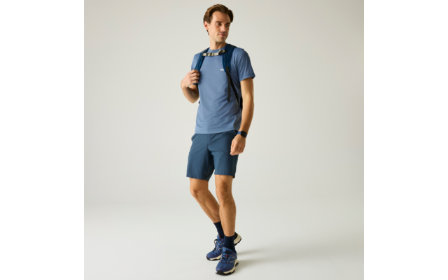 Ein Mann steht, trägt blaues T-Shirt, dunkelblaue Shorts, Rucksack mit Brustgurt, Sportschuhe und Smartwatch