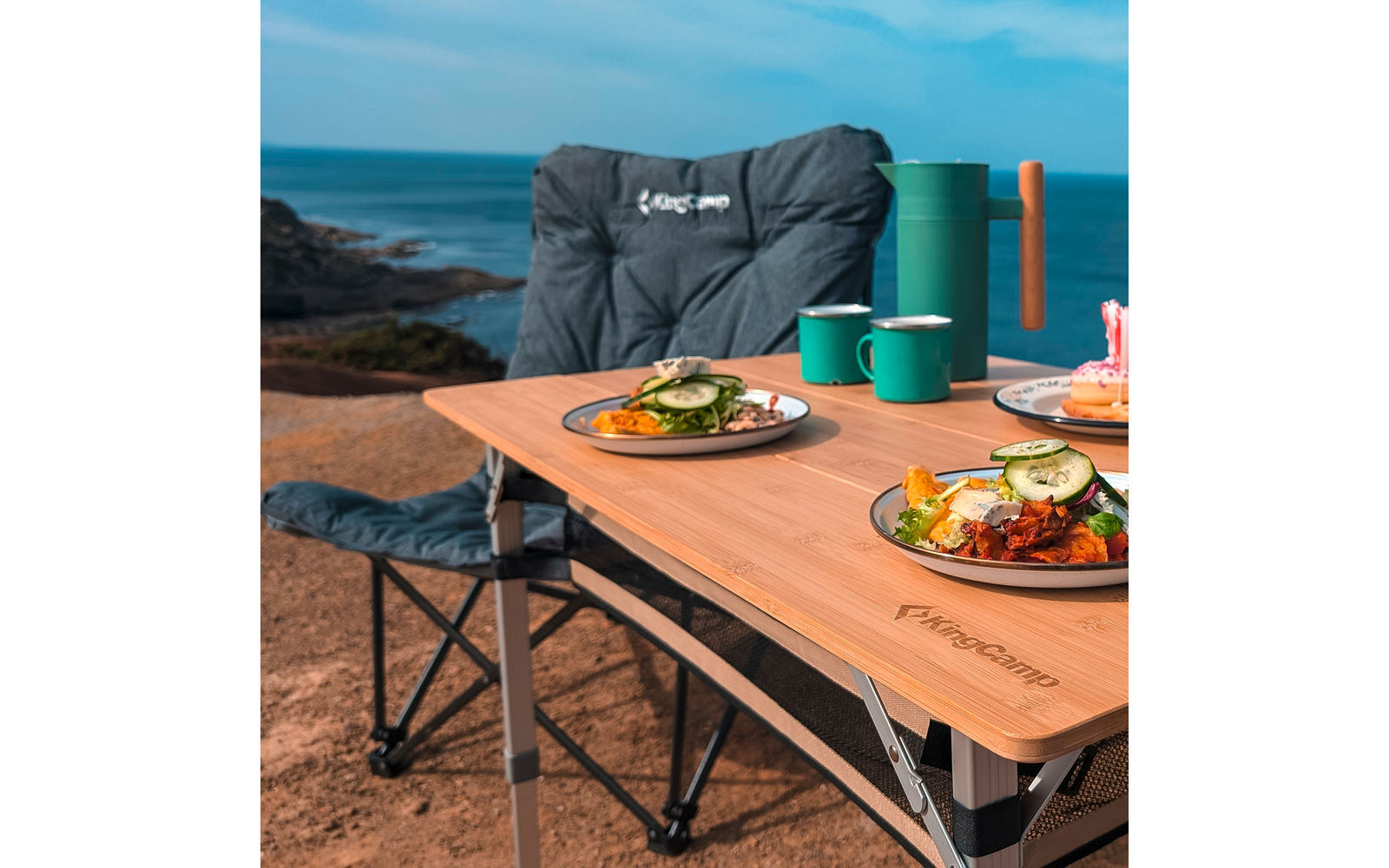 Ein Campingstuhl neben einem Holztisch mit zwei Tellern Essen und zwei Tassen am Meer