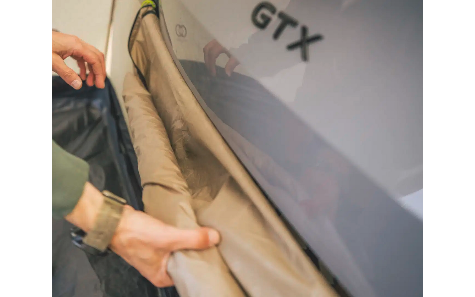 Hand zieht beige Hose aus, grau-schwarzes Kleidungsstück mit Aufdruck GTX im Hintergrund