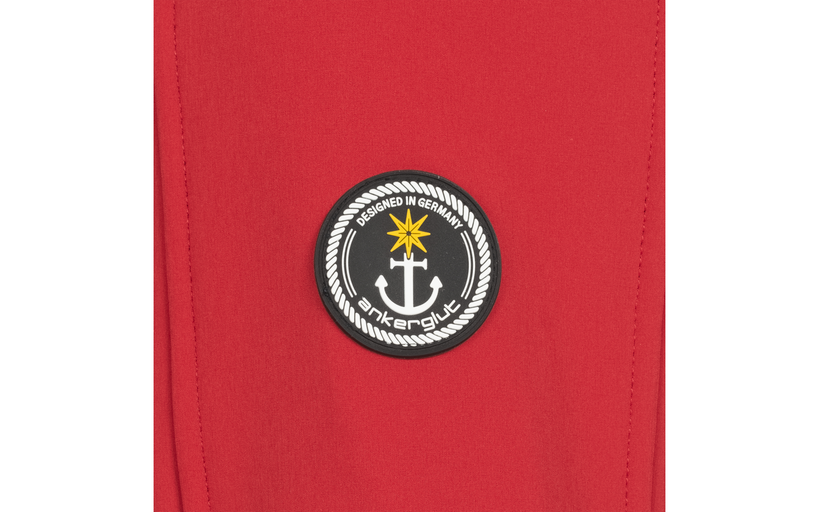 Roter Stoff mit rundem schwarzem Aufnäher. Aufnäher zeigt einen weißen Anker, einen gelben Stern oben, eine weiße Seilrand‑Umrandung und weiße Schrift DESIGNED IN GERMANY sowie ankerglut