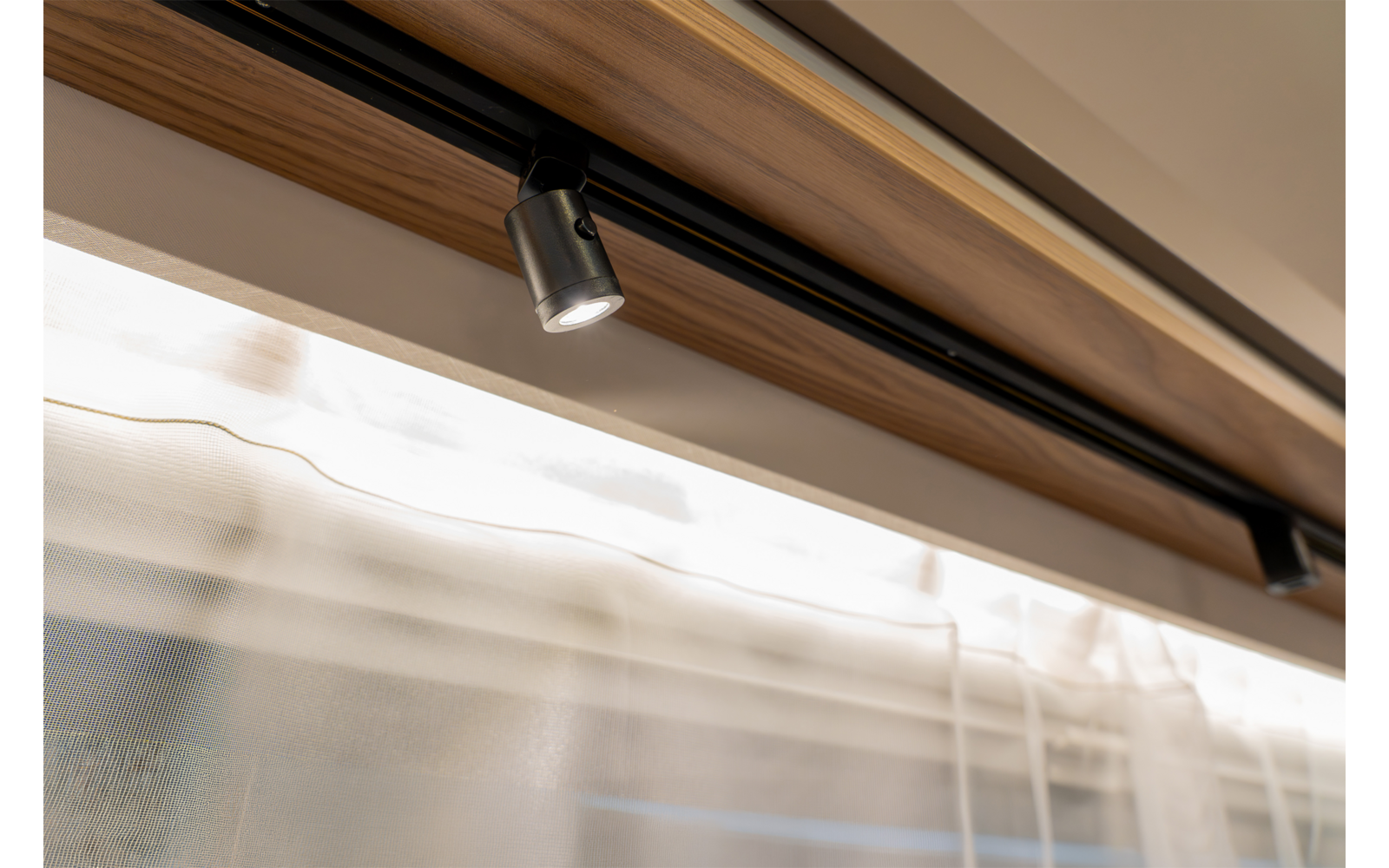 Schwarze Schienenleuchte mit einem verstellbaren Spot an einer Holzleiste an der Decke, darunter eine weiße transparente Gardine vor einem Fenster