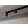 Schwarze Schiene mit einem Spot an einer hellen Holzdecke über einer strukturierten hellgrauen Wand