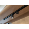 Schwarze Schienenleuchte mit einem eingeschalteten Strahler an einer hellen Holzdecke