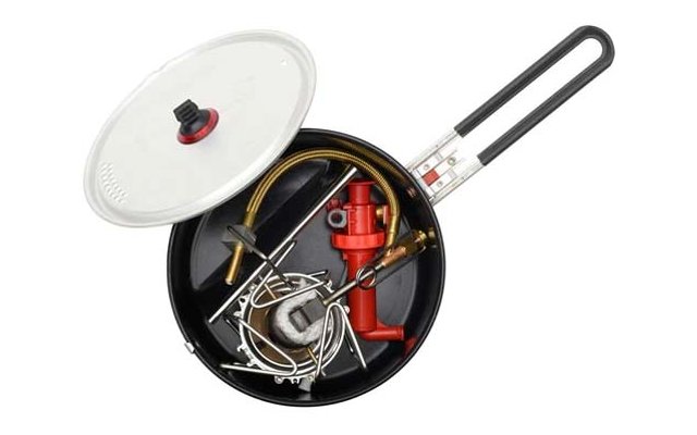 Schwarzer Topf mit langem Griff und transparentem Glasdeckel; im Inneren verschiedene Metallteile: zylindrisches rotes Metallteil, gelb ummantelter Schlauch, Metallstäbe und Drahtbügel