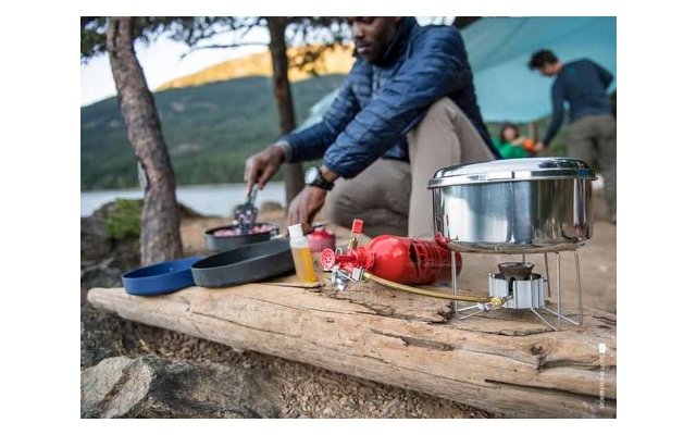 Person in blauer Jacke rührt in einer Pfanne auf einem Holzstamm; im Vordergrund Campingkocher mit roter Brennstoffflasche, Metalltopf und Pfannen; im Hintergrund Zelt, zwei Personen, See und bewaldete Hügel