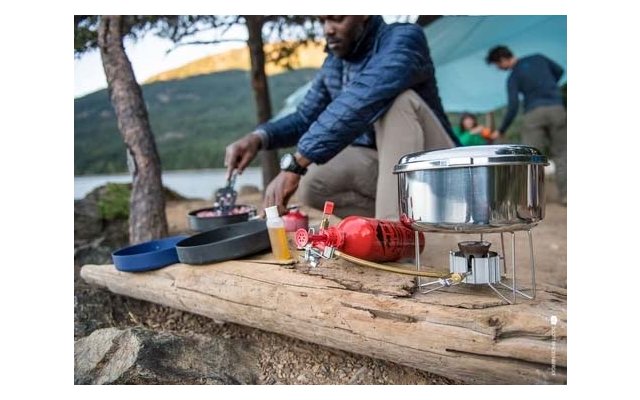 Person in blauer Jacke rührt in einer Pfanne auf einem Holzstamm; im Vordergrund Campingkocher mit roter Brennstoffflasche, Metalltopf und Pfannen; im Hintergrund Zelt, zwei Personen, See und bewaldete Hügel