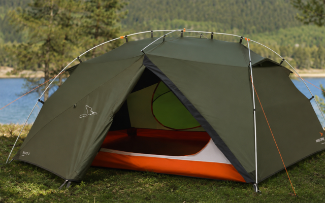 Green dome tent with open front flap showing orange-and-white interior, pitched on grass by a body of water with trees and hills in the background; tent poles and guy lines visible