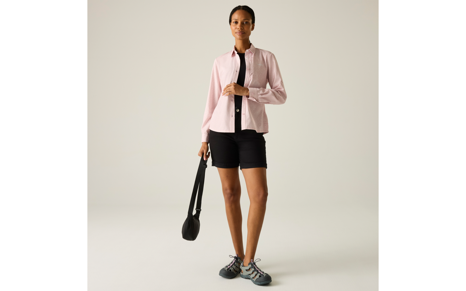 Stehende Frau in rosa Hemd über schwarzem Top und schwarzen Shorts, hält eine schwarze Umhängetasche in der linken Hand, trägt graue Sportschuhe, heller einfarbiger Hintergrund