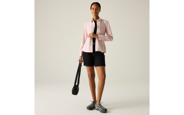 Person steht vor hellem Hintergrund, trägt hellrosa Hemd über schwarzem Top, schwarze Shorts, graue Sneaker und hält eine schwarze Tasche in einer Hand