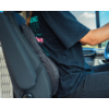 Person seated side-on at a vehicle's steering wheel, wearing a black T‑shirt and black pants, grey lumbar cushion attached to the driver's seat, car window with blue sky visible