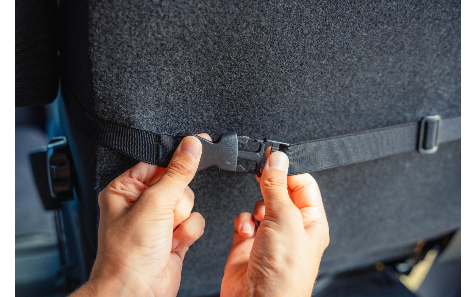 Two hands fasten a black plastic buckle on a strap attached to the back of a car seat