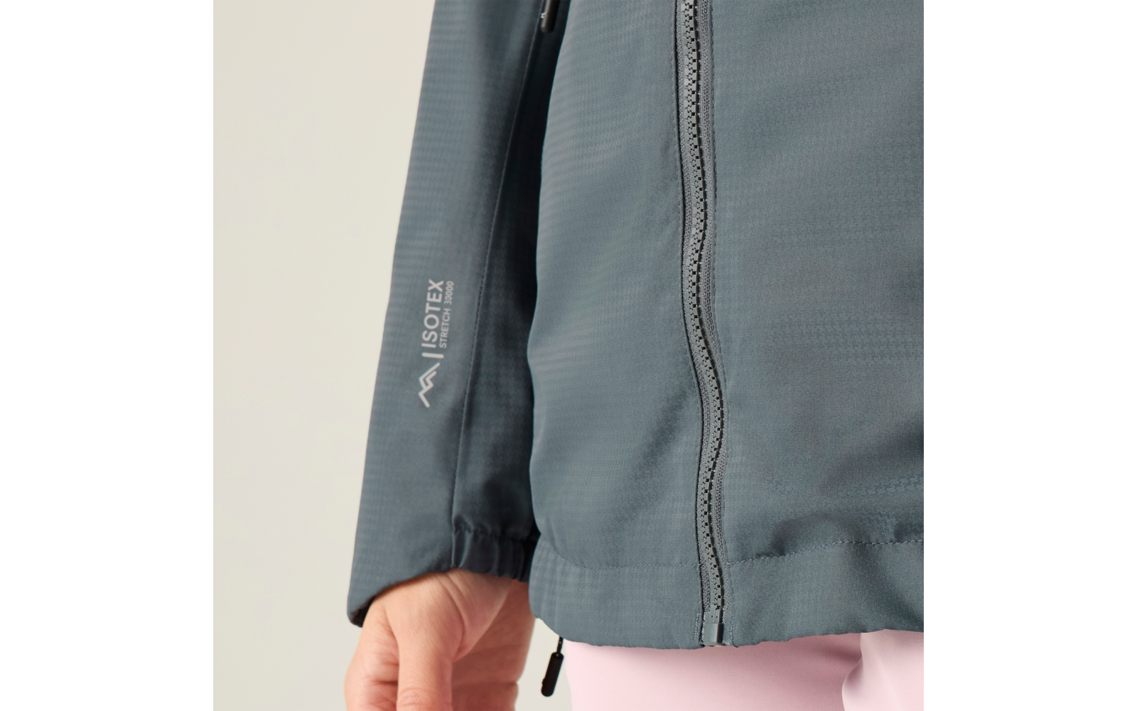 Nahaufnahme eines Oberkörpers in blaugrüner Jacke mit sichtbarem Reißverschluss, ISOTEX-Logo am Ärmel, elastischem Ärmelbund, sichtbarer Hand und rosa Hose