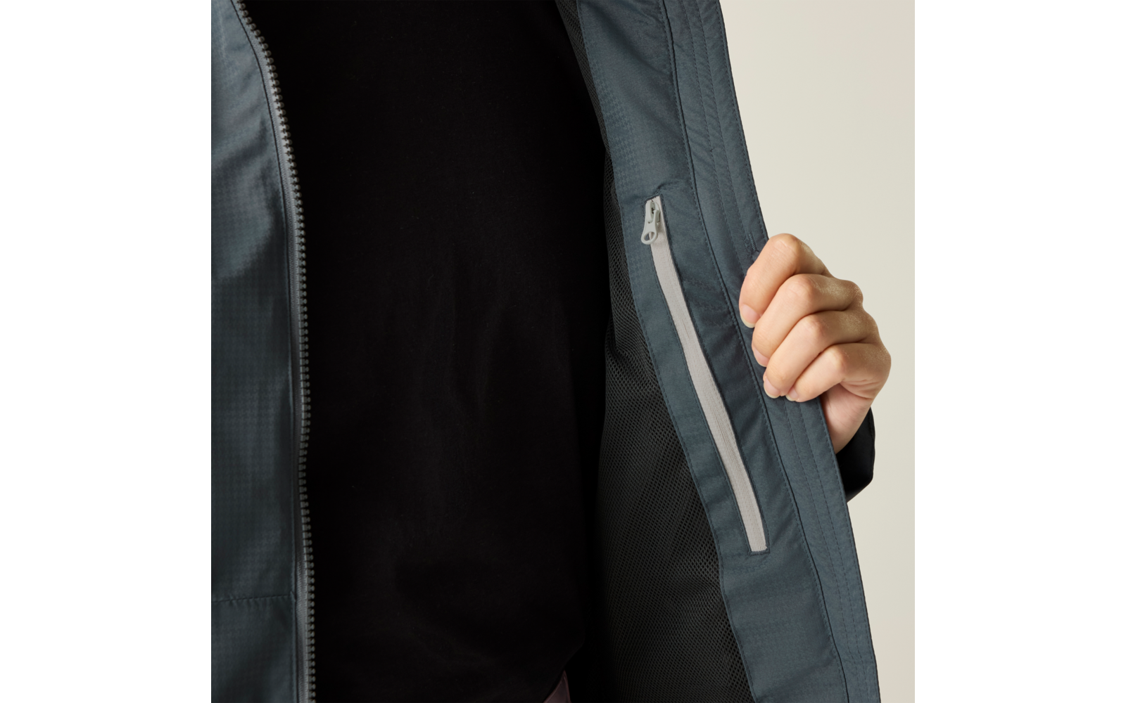 Person mit schwarzem T-Shirt hält offene graue Jacke, Innenseite mit senkrechter Reißverschlusstasche sichtbar, heller Hintergrund