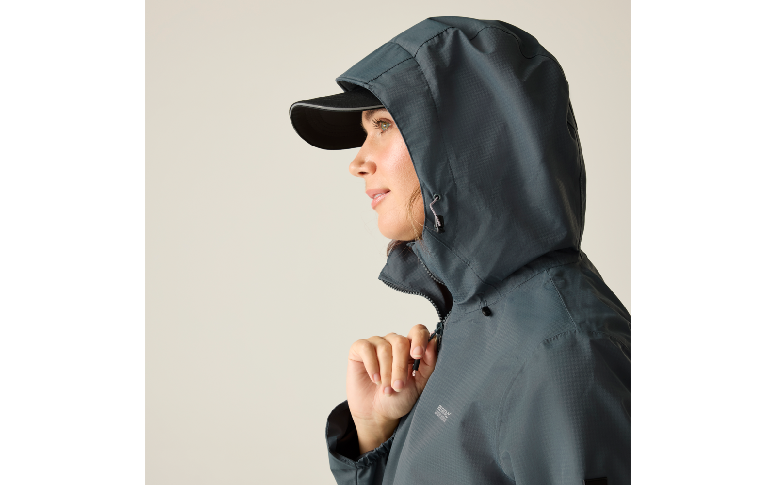Frau im Profil mit dunkler Regenjacke und Kapuze, trägt eine Schirmmütze, Hand an der Reißverschlussleiste