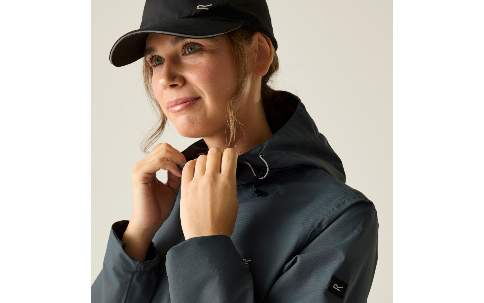 Person mit schwarzer Kappe und dunkler Kapuzenjacke, hält den Jackenkragen mit beiden Händen, schaut zur Seite, heller einfarbiger Hintergrund