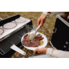 Hände halten einen weißen Teller mit Steak, mehreren Würstchen und Senf; eine Hand hält eine Grillzange über einem Grill