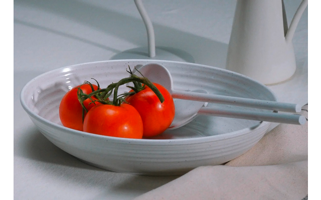 Cuenco poco profundo de cerámica blanca con tres tomates rojos en rama, cuchara de servir blanca, paño claro y jarra blanca al fondo