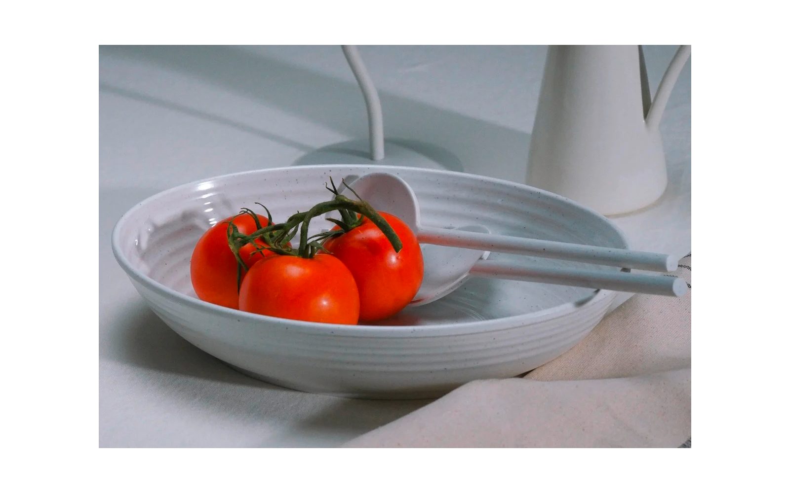 Cuenco poco profundo de cerámica blanca con tres tomates rojos en rama, cuchara de servir blanca, paño claro y jarra blanca al fondo