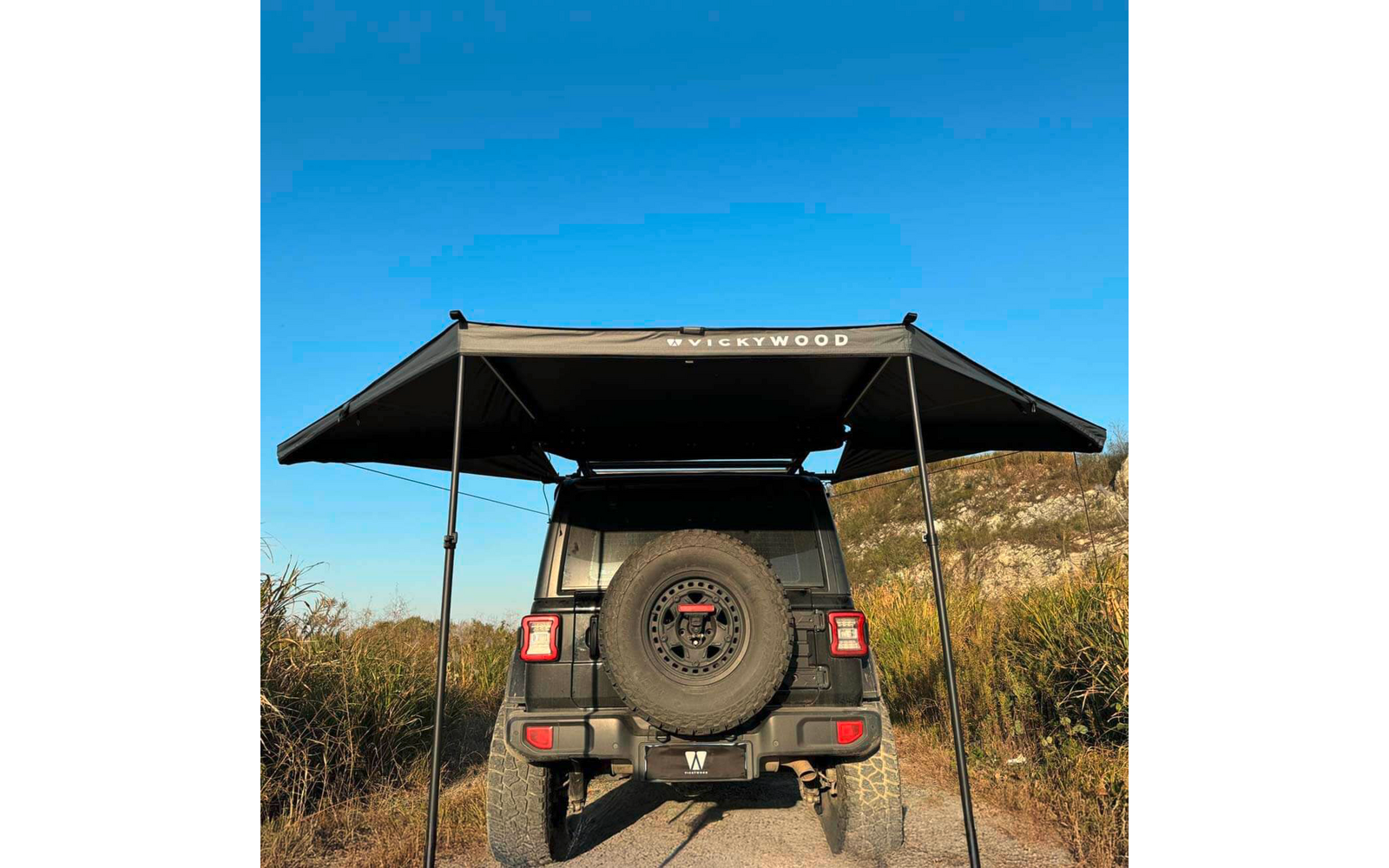 Heckansicht eines dunklen Geländewagens mit montiertem Reserverad und ausgefahrener Dachmarkise, zwei Stützstangen, Schotterweg, Gras, blauer Himmel