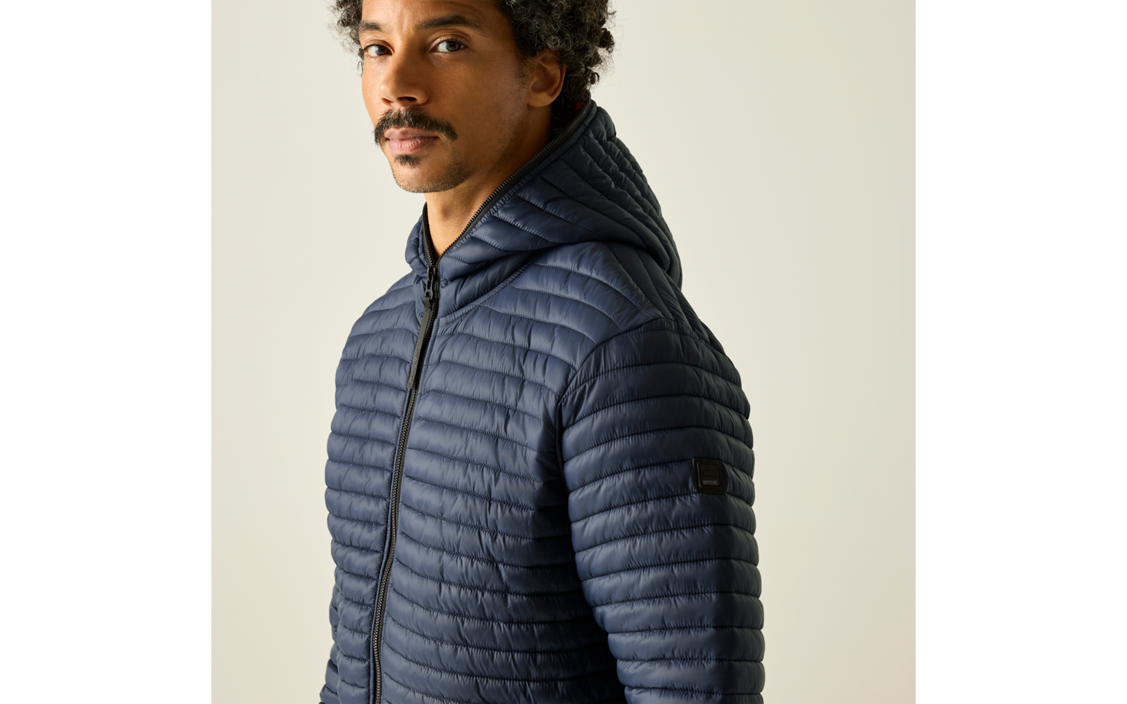 Person im Halbporträt trägt dunkelblaue gesteppte Kapuzenjacke, kurze lockige Haare und Schnurrbart, blickt leicht zur Seite, heller Hintergrund