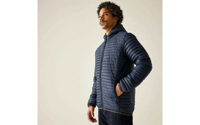 Person mit kurzen dunklen lockigen Haaren und Schnurrbart, trägt eine dunkelblaue gesteppte Jacke mit Kapuze, steht vor hellem einfarbigem Hintergrund und blickt nach oben rechts