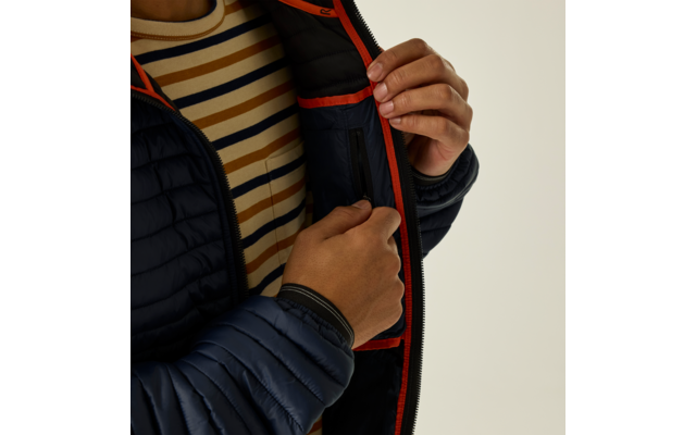 Oberkörper einer Person in gestreiftem T‑Shirt, die eine dunkelblaue gesteppte Jacke mit orange-rotem Reißverschlussrand öffnet und eine Innentasche zeigt