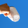 Hand hält einen weißen, facettierten Becher vor blauem Himmel