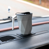 Grau facettierter Becher mit Henkel und schwarzem Deckel auf dem Armaturenbrett eines Autos, Lüftungsgitter und unscharfe Straße durch die Windschutzscheibe sichtbar