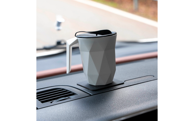 Grau facettierter Becher mit Henkel und schwarzem Deckel auf dem Armaturenbrett eines Autos, Lüftungsgitter und unscharfe Straße durch die Windschutzscheibe sichtbar