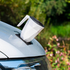 Weiße, facettierte Tasse mit schwarzem Deckel und Henkel auf einer schwarzen Matte auf der Motorhaube eines silbernen Autos; unscharfer grüner Hintergrund