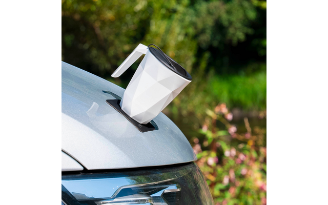 Weiße, facettierte Tasse mit schwarzem Deckel und Henkel auf einer schwarzen Matte auf der Motorhaube eines silbernen Autos; unscharfer grüner Hintergrund