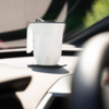 Weißer Becher mit facettiertem Muster und schwarzem Deckel auf einer schwarzen Haftunterlage auf dem Armaturenbrett eines Autos, verschwommene Windschutzscheibe im Hintergrund