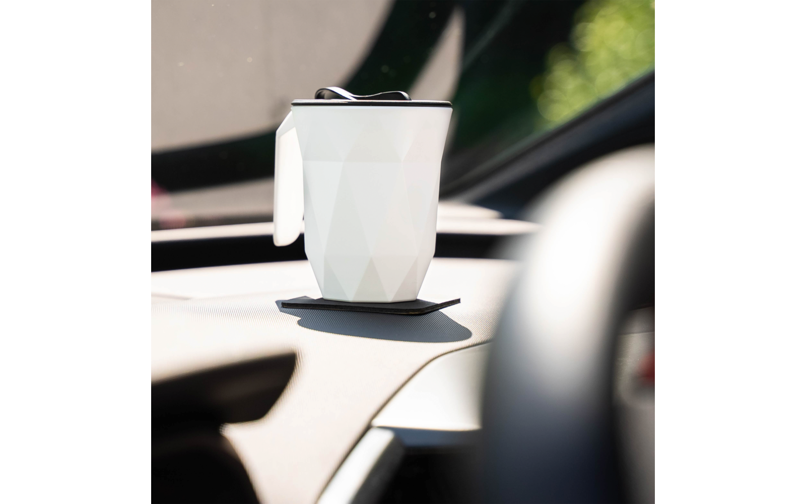 Weißer Becher mit facettiertem Muster und schwarzem Deckel auf einer schwarzen Haftunterlage auf dem Armaturenbrett eines Autos, verschwommene Windschutzscheibe im Hintergrund