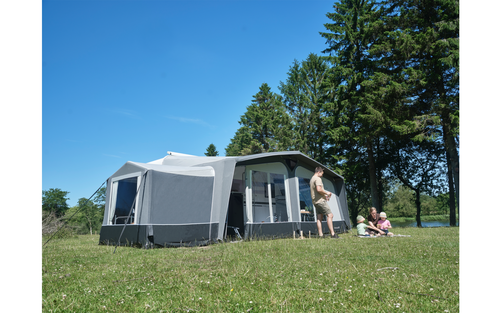 Großes graues Camping-Vorzelt auf einer Wiese, eine erwachsene Person steht davor, zwei Kinder sitzen auf einer Decke, im Hintergrund Bäume und blauer Himmel