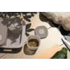 Beige bewaarbakjes en deksel, een open klein bakje met zaden, een mes, een houten snijplank en een groene courgette op een lichte tafel, bladschaduwen bovenaan