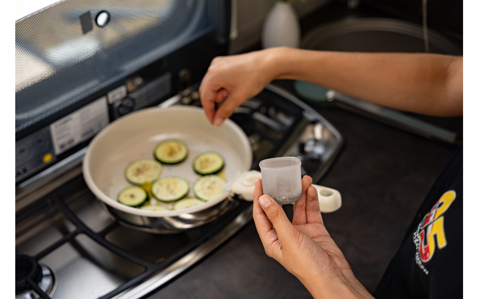 Handen strooien kruiden uit een klein bakje over courgetteplakjes in een witte koekenpan op een gasfornuis; een lepel ligt in de pan