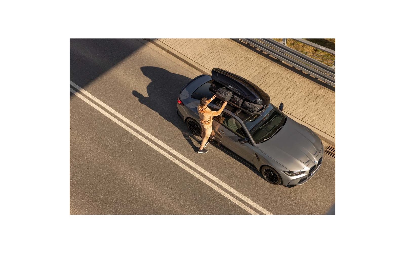 Vogelperspektive auf graues Auto am Straßenrand, eine Person in beiger Kleidung öffnet eine Dachbox